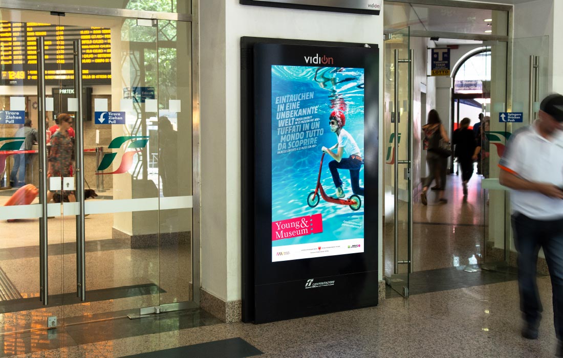 Digitales Plakat am Screen Vidion Digimupi der Centostazioni am Bahnhof Bozen. Young & Museum Kampagne mit einer Grafik von Waldemar Kerschbaumer, Grafikbüro adpassion Südtirol.